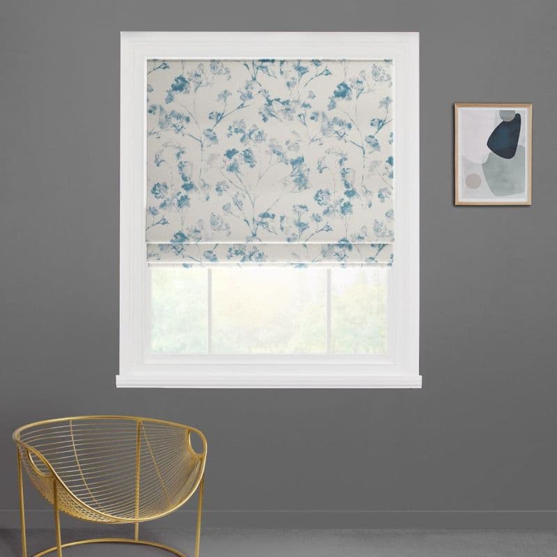 Floral Roman blind, blue botanical print, partially lowered and filtering daylight over a white-framed window; gray-walled interior with a gold wire chair and a small abstract framed print nearby.