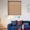 Honey‑tan pleated cellular blind, lowered and closed with fine horizontal ridges and a slight bottom gap, filtering daylight; modern living room with blue sofa, colored cushions, floor lamp and sideboard.