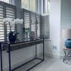 White plantation shutters covering lower window panels, angled louvers partially open, filtering soft daylight; in front, a slim metal-framed console table with orchid and decor, pale-carpeted modern sitting area.