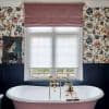 Pink Roman blind folded at top; white pleated blinds lowered, diffusing daylight over a double-sash window above a freestanding pink bathtub with dark wainscoting and floral wallpaper.