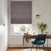 Patterned mauve Roman blind lowered across the window, partially blocking light; a tidy home-office desk beneath holds a mirror, scarf, vase of flowers and a teal chair against neutral walls.