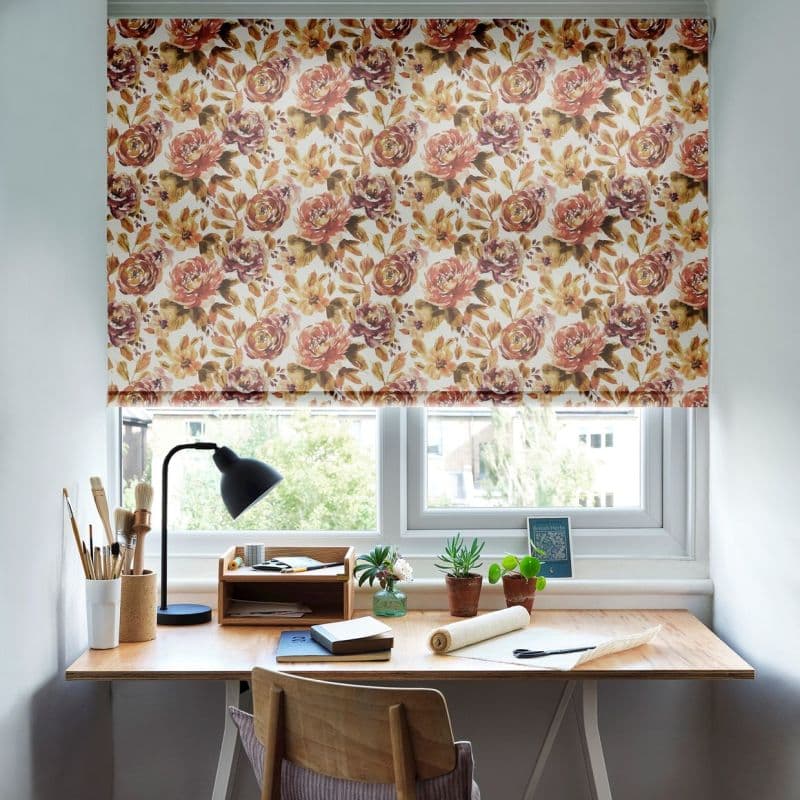 Floral roller blind with peach-and-ochre roses lowered across the window, filtering daylight; a tidy wooden desk beneath with lamp, potted succulents, brushes and notebooks in a calm home workspace.