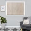 Beige patterned vertical blinds, scalloped bottoms, fitted inside a white frame — closed, softly filtering daylight. Context: grey-walled living room with armchair, cushion, potted plant and framed print.