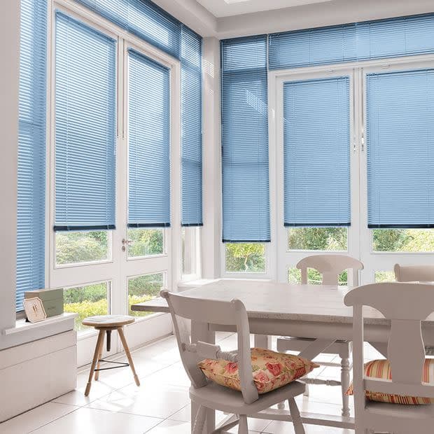 Light-blue horizontal Venetian blinds covering tall bay windows, lowered and filtering soft daylight with small bottom gaps; white dining table and chairs in a bright sunroom overlooking a green garden.