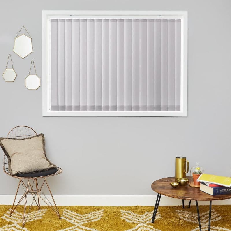 Vertical blinds, closed and evenly spaced across a white-framed window, softly diffusing daylight; modern living room with gray walls, gold-accent decor, wire chair, round wooden coffee table and mustard rug.