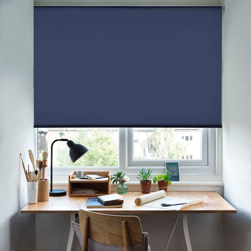 Navy blue roller blind, smooth fabric with straight hem, lowered across the window, blocking most daylight with a narrow gap; home workspace with wooden desk, lamp, potted plants, notebooks, chair, daylight outside.