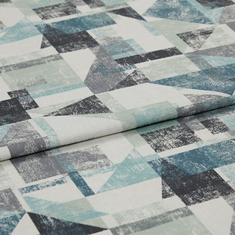 Upholstery fabric (distressed geometric teal, grey, black triangles and squares) folded with overlapping crease; close-up on a flat surface in neutral studio lighting, no visible surroundings.