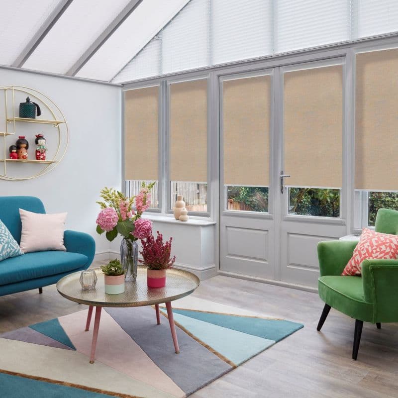 Beige roller blinds, lowered across the upper panes of tall glazed doors and windows, filtering daylight; conservatory-style living room with colorful seating, coffee table, and plants.