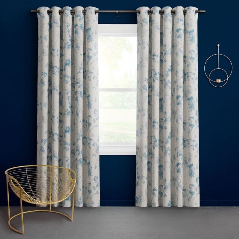 Grommet-top white curtains with blue floral print, hung on a metal rod and slightly parted, framing a bright window; navy-blue wall, gold wire chair and circular wall sconce, daylight.