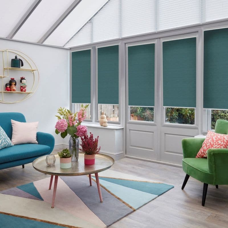 Teal roller blinds lower over upper panes of tall windows and French doors, blocking light; bright conservatory-style living room with vaulted glass roof, colorful sofas, coffee table and plants.