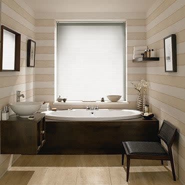 White pleated cellular blind fully lowered over window, softly diffusing daylight; a built-in bathtub beneath with dark wood paneling, vessel sink, striped beige walls, wooden floor, minimal shelf.