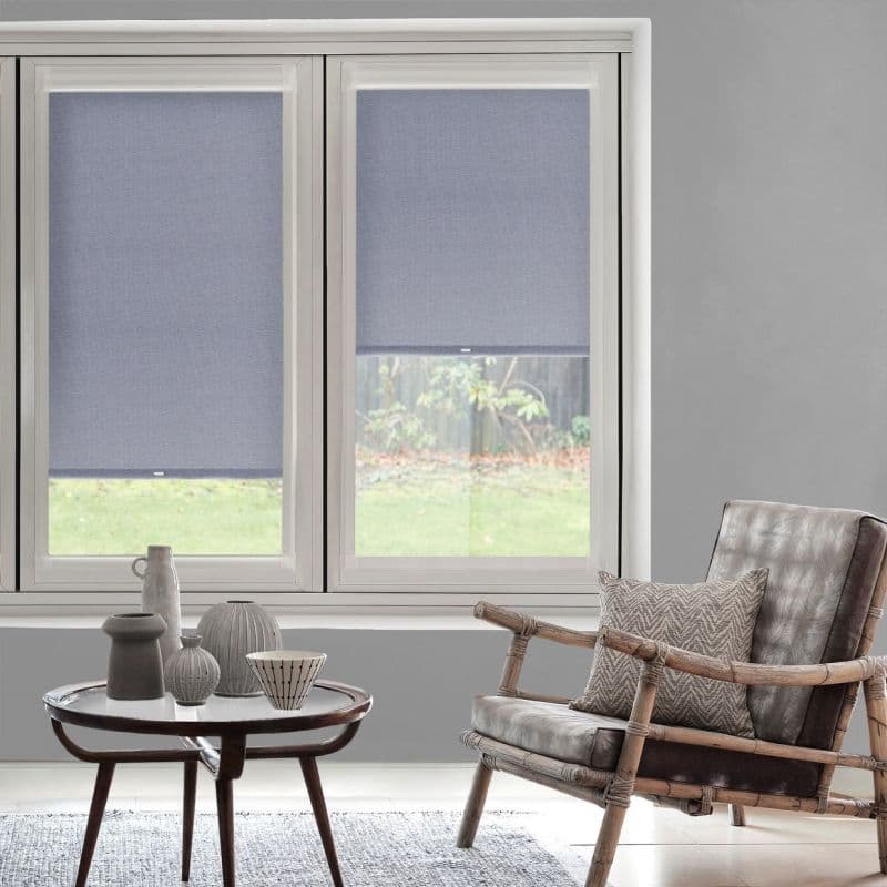 Two blue-gray roller blinds, partially lowered and filtering daylight, cover twin windows in a gray living room with wooden-framed leather armchair, cushion, round coffee table and ceramic vases; lawn outside.