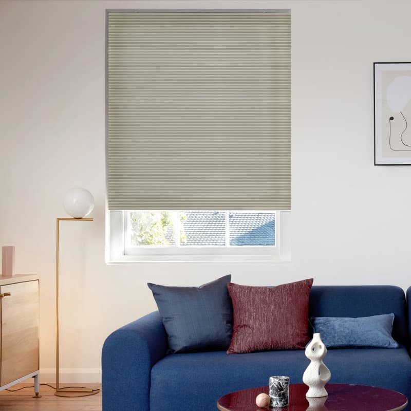 A beige cellular blind pulled down over a recessed rectangular window, closed and filtering daylight into a modern living room with a blue sofa, cushions, floor lamp, and framed art.