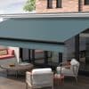Turquoise retractable fabric awning extended over patio—mounted above large sliding glass doors, shading outdoor seating with rope-accent armchairs, round wooden coffee table, brick upper wall and daylight view.