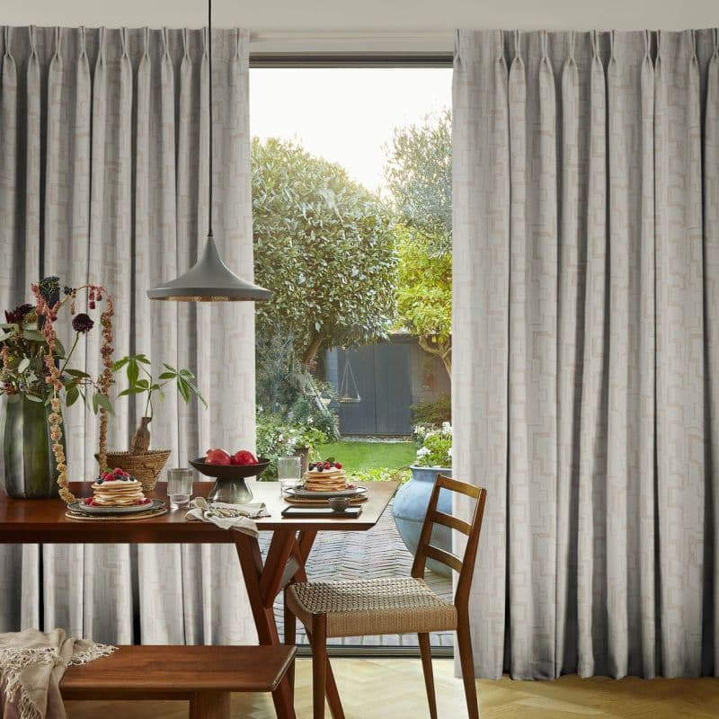 Pleated beige curtains, full-length and parted centrally, revealing a sliding glass door; framing and filtering daylight into a dining area with a wooden breakfast table and a lush garden beyond.