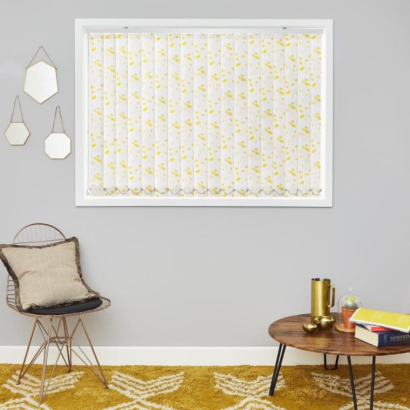 Sheer vertical blinds with yellow leaf pattern, fully closed and gently filtering daylight, set in a white-framed window above a gray-walled living area with wire chair, cushion, wooden coffee table, mustard rug.