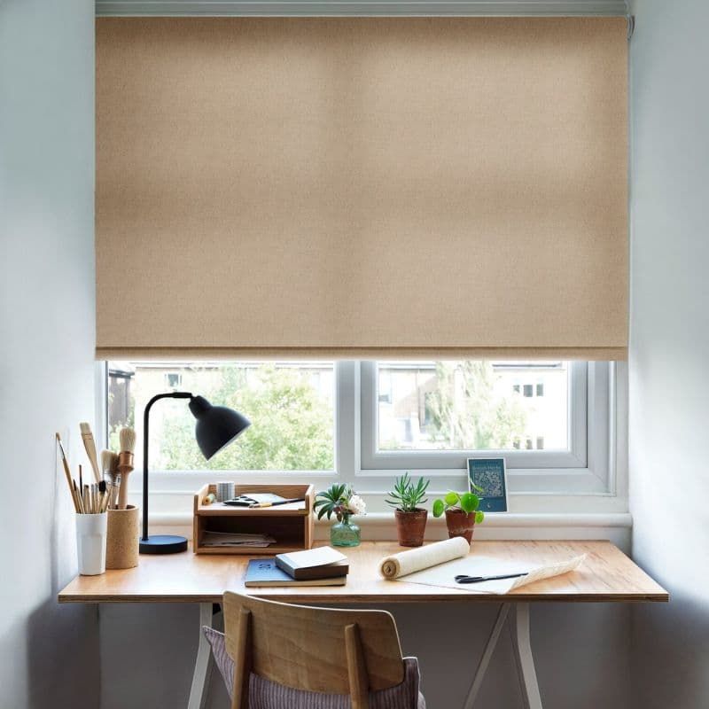Beige roller blind, fully lowered with visible top roller and textured fabric; blocking and softly filtering daylight; above a wooden desk with lamp, brushes, potted plants and a green view.