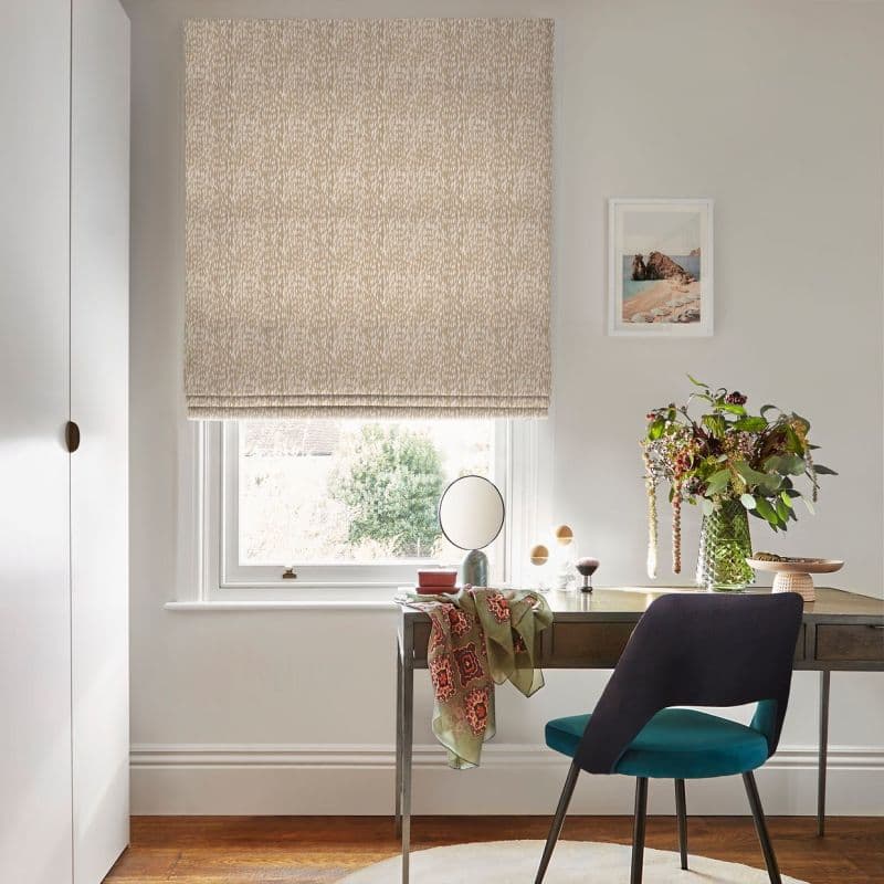 A patterned beige Roman blind, fully lowered with soft folds, filtering daylight; covering a sash window above a tidy desk with a chair, vase of flowers and framed wall art.