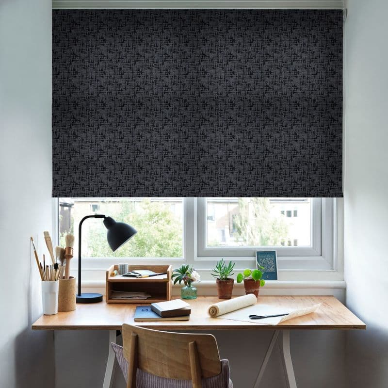 Dark charcoal patterned roller blind, lowered to cover a double window and block light; wooden desk beneath with lamp, brushes, notebooks and potted plants in a bright home office.
