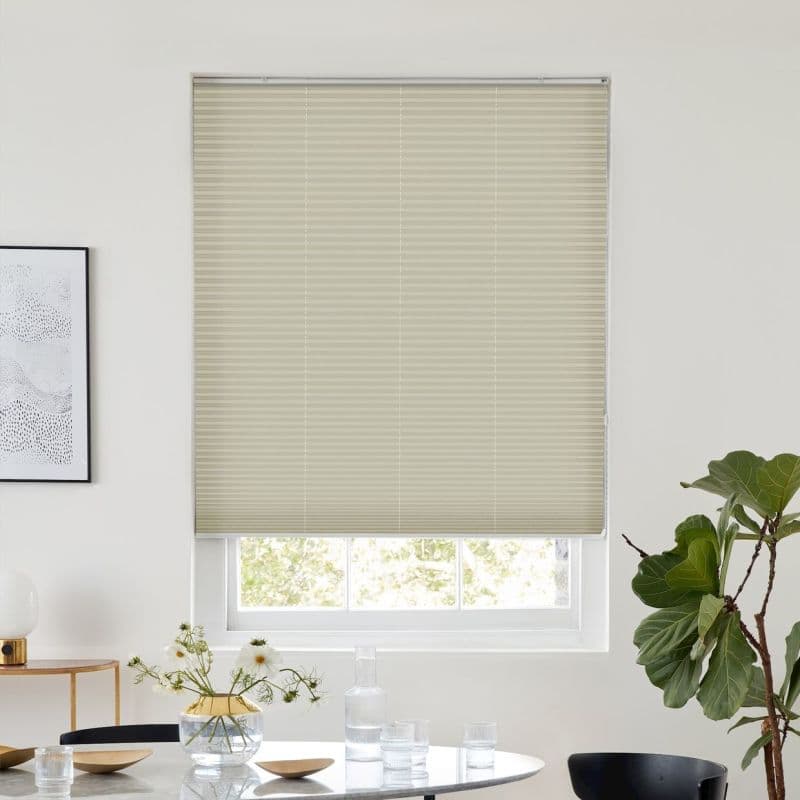 Light-beige cellular blind, fully lowered with horizontal pleats, filtering daylight; modern dining room with round table, vase and glassware, potted fiddle‑leaf fig visible by the window.