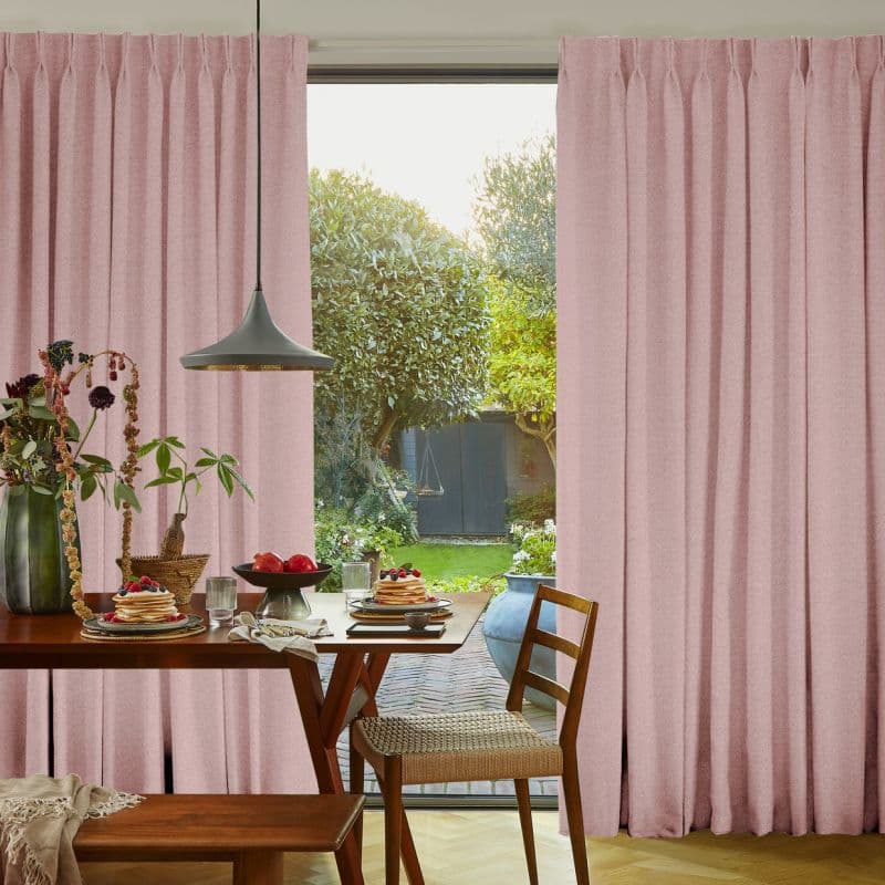 Pleated blush-pink floor-to-ceiling curtains, slightly parted and filtering light, frame a breakfast table with wooden chairs and pendant lamp, opening onto a sunlit garden.