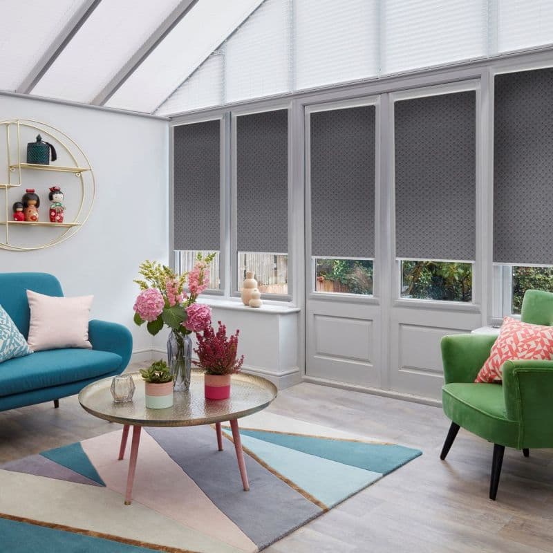 Grey patterned roller blinds covering tall doors and windows, lowered mid-height and filtering daylight; bright conservatory living room with teal sofa, green armchair, floral centerpiece and geometric rug.