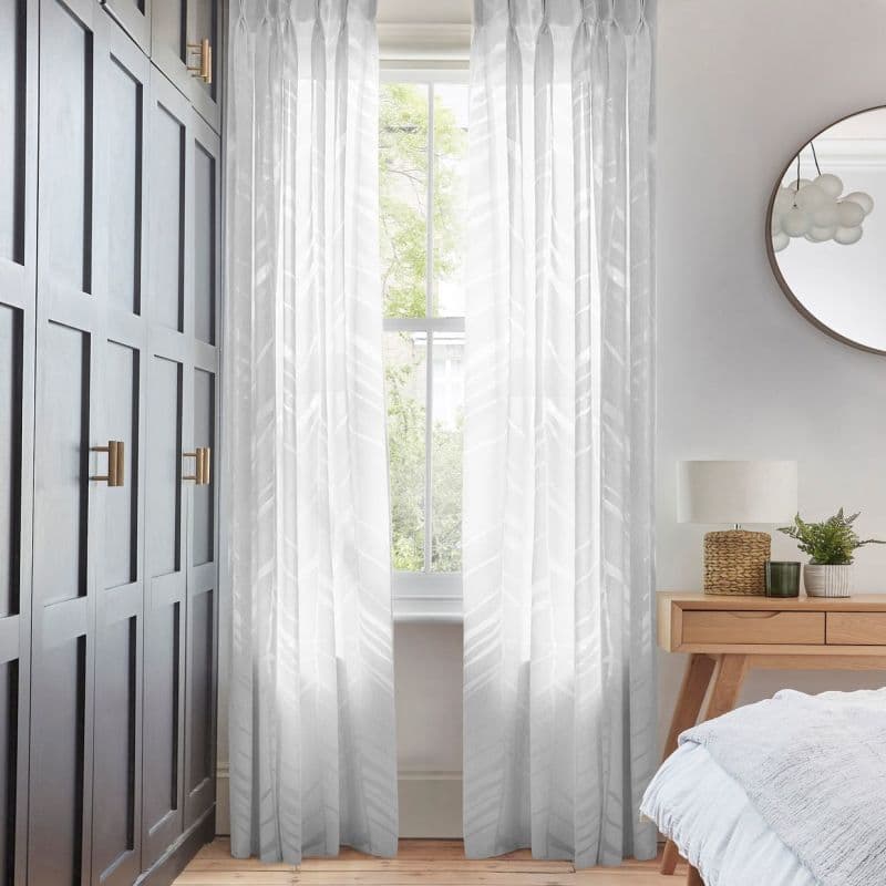 Sheer patterned curtains framing a tall sash window, filtering soft daylight into a bedroom corner with dark paneled wardrobe, wooden bedside table, lamp, mirror, and green outside view.