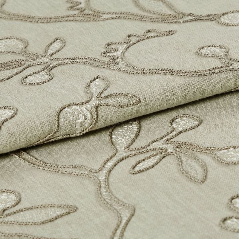 Beige embroidered fabric folded into overlapping layers, showing raised stitched vine-and-leaf motifs in tonal thread; close-up view with soft, even lighting emphasizing texture.