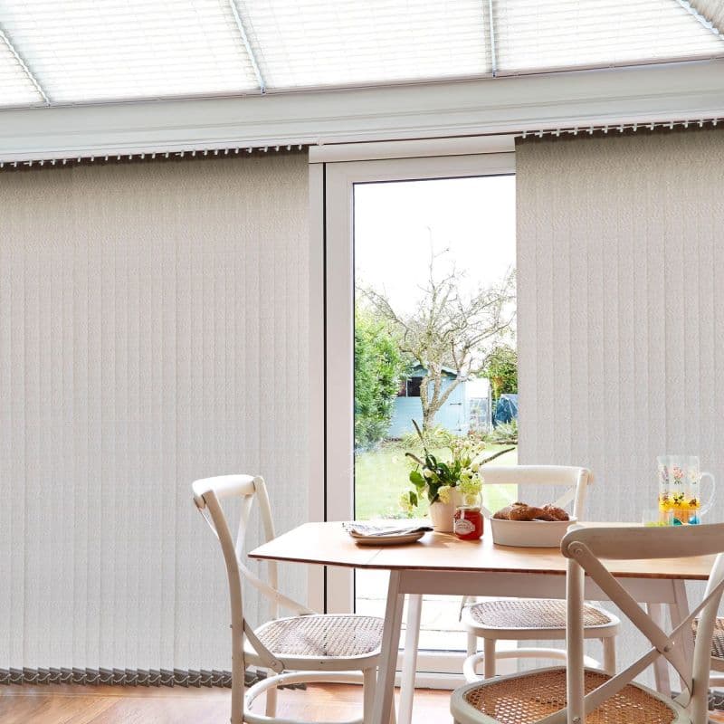 Beige vertical blinds with slats cover a sliding door; central gap reveals outdoors. Partially open, they filter daylight into a bright dining nook with table, chairs, vase, garden view.