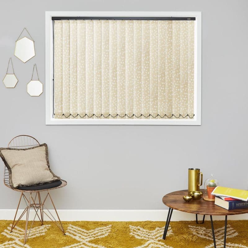 Cream vertical blinds with white leaf pattern, hanging closed across a window with beaded chain; filtering soft light in a gray living room with chair, mustard rug and side table.