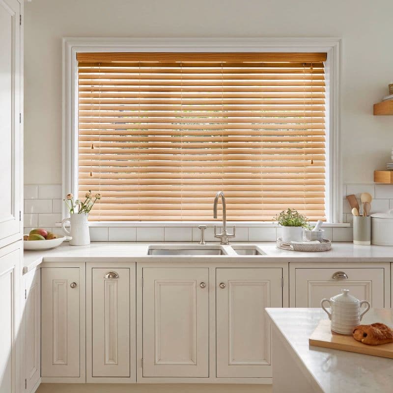 Wooden horizontal blinds across a large window, lowered with slats partly angled, filtering warm daylight above a white kitchen sink and countertop with cabinets, potted plant, utensils, teapot and croissant.