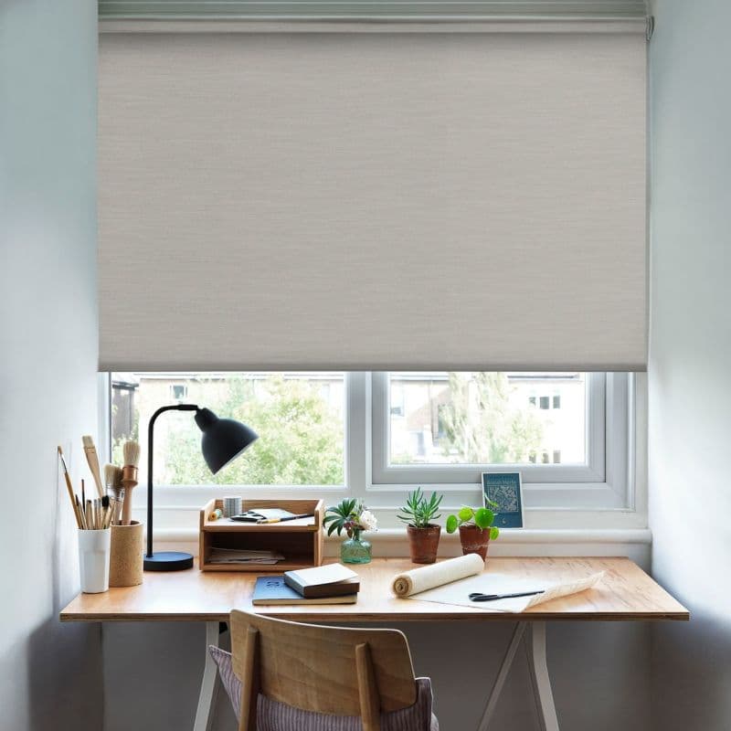 Light beige roller blind lowered across the window, filtering daylight; small wooden desk beneath with lamp, art brushes, notebooks and potted succulents in a bright workspace.
