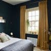 Floor-length mustard velvet pleated curtains, hung on a rod and pulled to the sides, slightly pooling on the floor; framing a sunlit bedroom window with plants, radiator, dark teal walls.