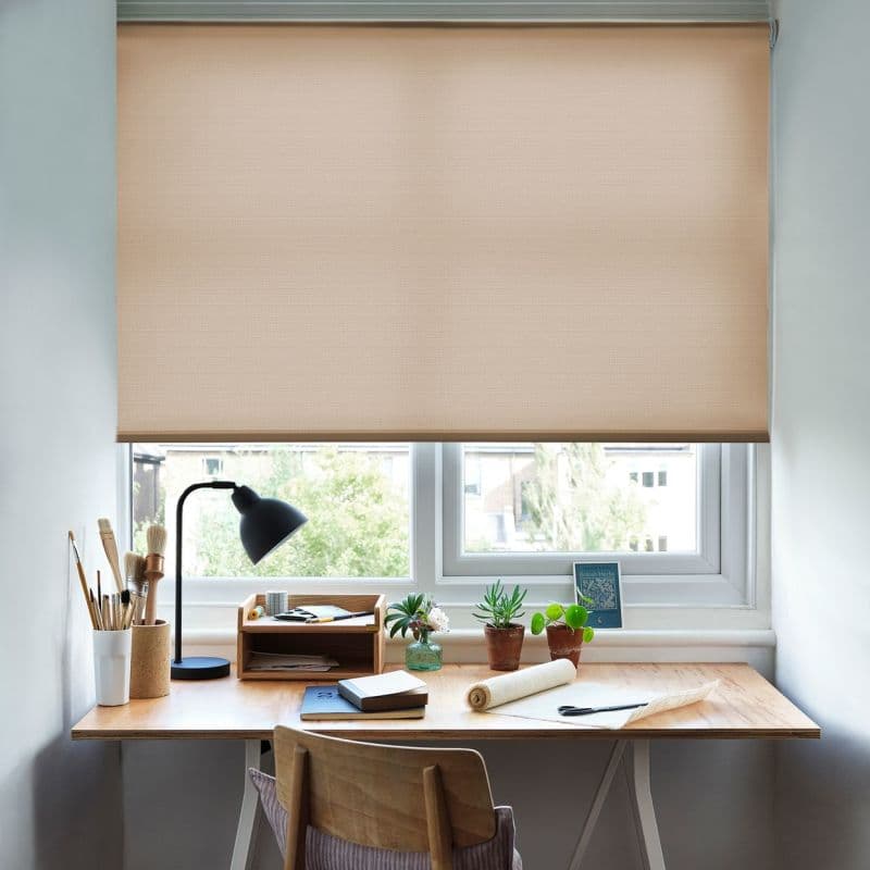 Beige roller blind, textured fabric, fully lowered across a double-pane window, filtering soft daylight; compact wooden desk beneath holds a lamp, art brushes, notebooks and potted plants, pale-walled room.