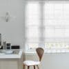 Sheer white Roman shade lowered over a multi-pane window, gently filtering daylight; minimalist workspace with white desk, wooden chair, pendant lamp and soft natural light revealing bare tree branches outside.