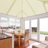 Cream pleated blinds cover the pitched conservatory roof plus windows and French doors, closed, filtering soft daylight; bright dining-kitchen space with wooden table, upholstered chairs, patio view.