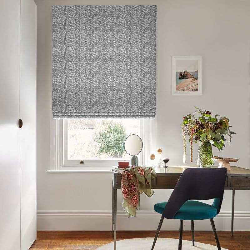 Grey patterned Roman blind lowered, softly diffusing daylight over a desk with mirror, vase of flowers and a draped scarf; teal chair on hardwood floor, pale walls and a small framed seascape.