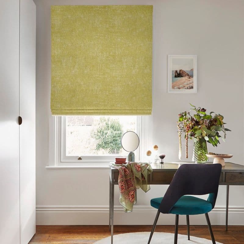 Textured chartreuse Roman blind, lowered with horizontal folds across a window; filtering daylight; above a wooden desk with teal chair, mirror, vase of flowers and coastal print in an office.