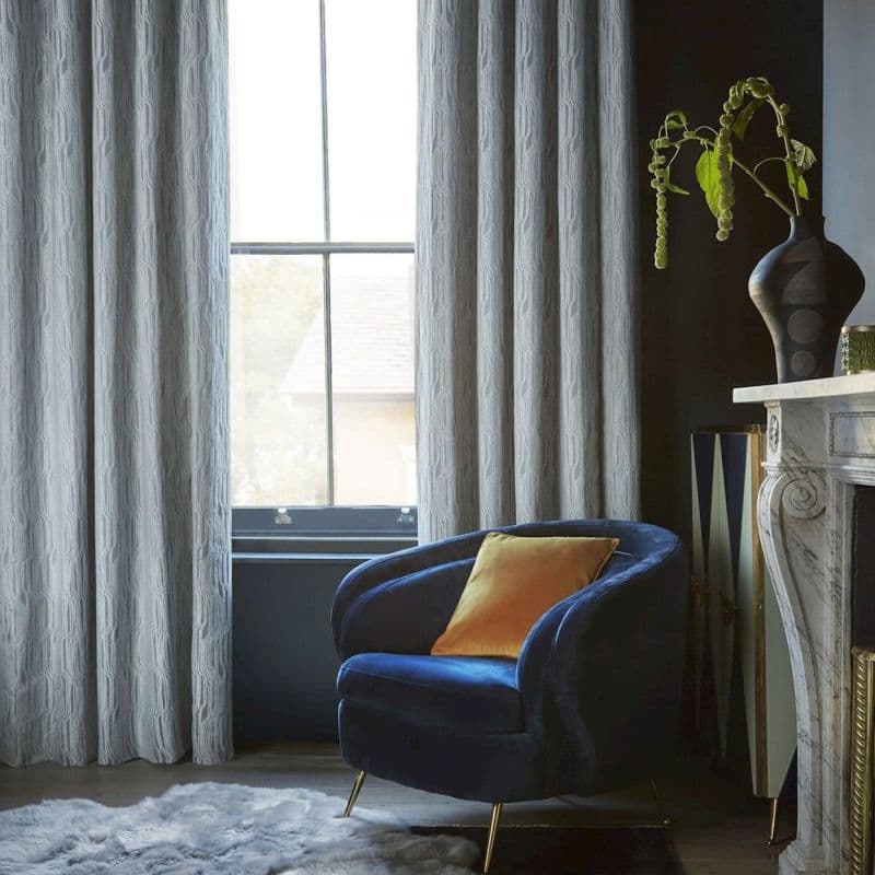Floor-to-ceiling textured gray curtains, hung in soft pleats framing a four-pane sash window, filtering pale daylight into a moody living room with a blue velvet armchair and marble mantel.