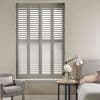 White four-panel louvered plantation shutters covering a tall rectangular window, slats tilted to filter daylight; bedroom setting with upholstered bed, side table with lamp, flowers and a tufted armchair.