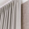 Pleated grey fabric curtains hung from a white ring-top rod, tightly closed and evenly pleated; they obscure a textured beige wall covering beside them, bathed in soft, diffused indoor light.