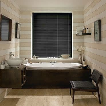 Black horizontal blinds, fully closed with thin slats covering a central window; blocking daylight; above a white tub in a warm wood-paneled bathroom with sink, shelf and chair.