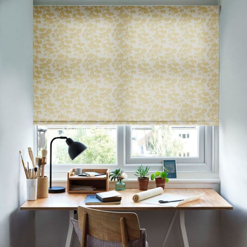 Yellow botanical-patterned roller blind fully lowered, filtering light; covering a double casement window. Below, a wooden desk in a bright home study holds a lamp, notebooks, potted plants, brushes.