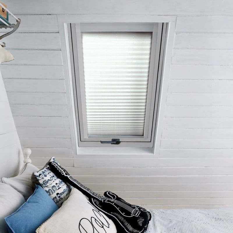 White pleated blind fully closed over a rectangular skylight, softly diffusing daylight; set in a bright white shiplap attic bedroom above a bed corner with blue, patterned and guitar-shaped cushions.
