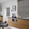 Horizontal aluminum Venetian blinds covering a large window at right and small door window at left, closed and filtering daylight; modern kitchen with light wood cabinets, black countertop, bar stools.