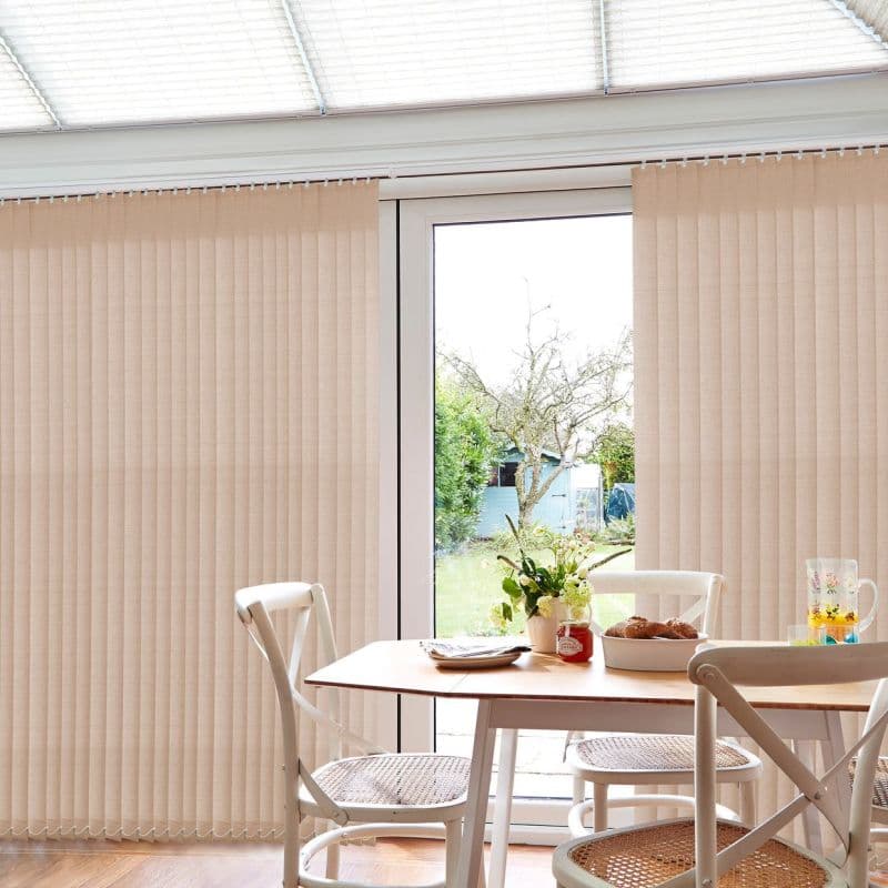 Beige vertical blinds covering floor-to-ceiling patio doors, parted centrally to reveal view; filtering soft daylight. Small wooden dining table with white woven chairs, vase and pastries, sunlit backyard beyond.