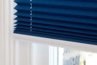 Deep-blue pleated (cellular) window blind, lowered and folded, with small white clip links along the left pleats; mounted inside a white frame, filtering soft daylight from outside.