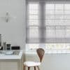 Sheer grey Roman blind, lowered with horizontal folds, filtering pale daylight over a multi‑pane window in minimalist home office with white desk, wooden chair, pendant lamp and bare trees outside.