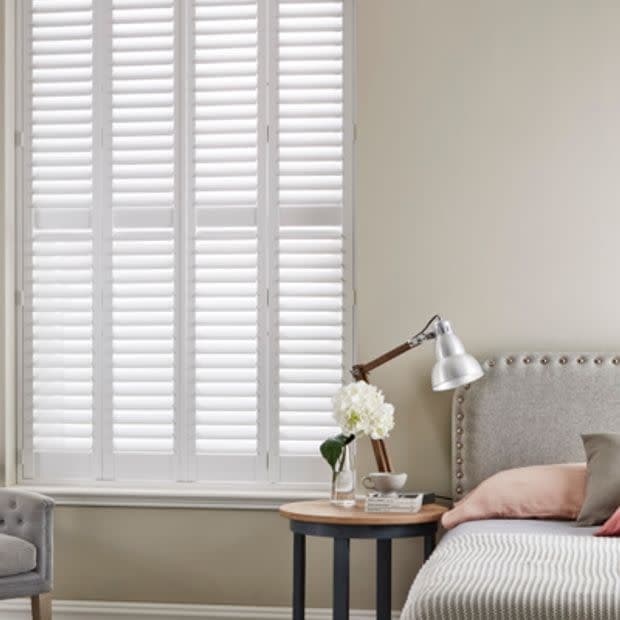 White plantation shutters covering a tall window, closed with slatted panels filtering soft daylight; beside a bedside table with lamp, vase, and upholstered headboard in a neutral bedroom.