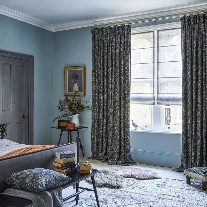 Patterned heavy curtains and sheer Roman blinds — curtains parted and pooling at the floor, blinds partially lowered filtering soft daylight — in a softly lit blue bedroom with rug, bench, and stacked books.